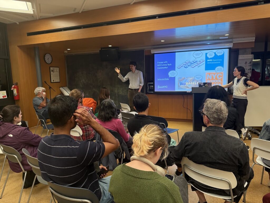 A community workshop in a library: one presenter gestures beside a slide about engaging with NYC’s civic tech community (logos for Open Data Week and NYC School of Data visible) while another stands at a podium; attendees sit in rows facing the screen.