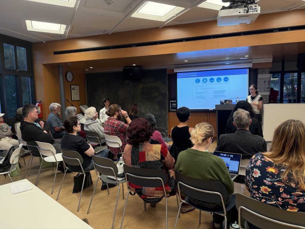 A presenter explains a “What is NYC Open Data?” slide showing icons and example datasets; about 15 attendees watch, including one person using a laptop in the front row.