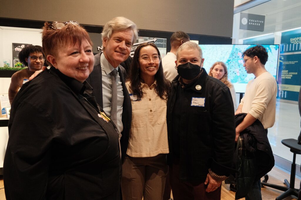 Group of six diverse adults posing for a photo at a tech event, with a large map display and glass-walled backdrop behind them.
