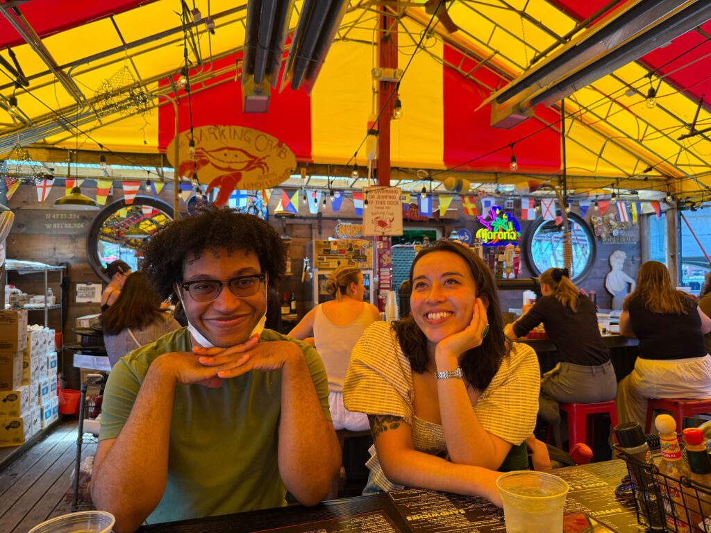 Two friends smile at the camera while leaning on a market stall counter under a bright yellow and red canopy.