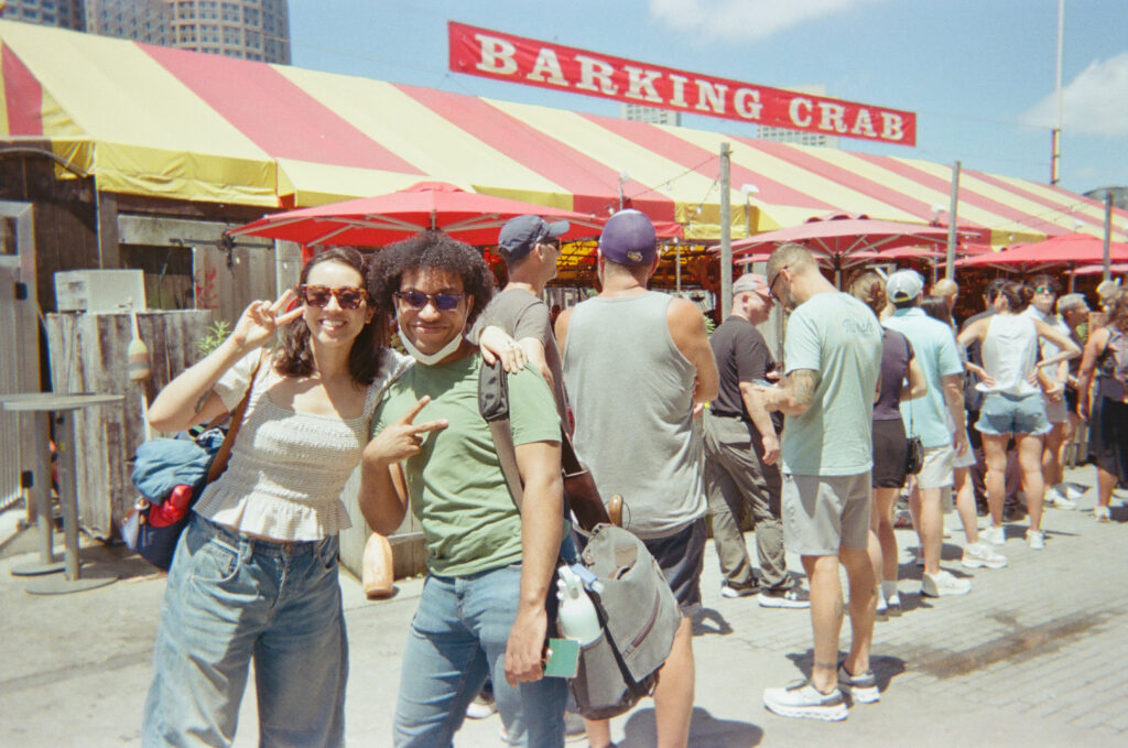 Two smiling people take a photo outdoors under a red-and-yellow canopy with the title "Barking Crab."