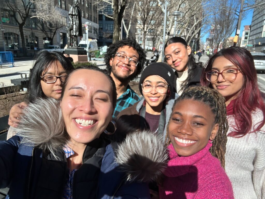 Group of smiling friends taking a selfie on a sunny city sidewalk in an urban area.