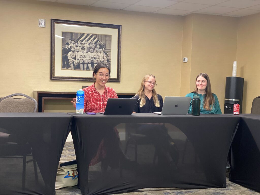 Three people sit at a table with their laptops and water bottles. The table is covered in a black cloth.