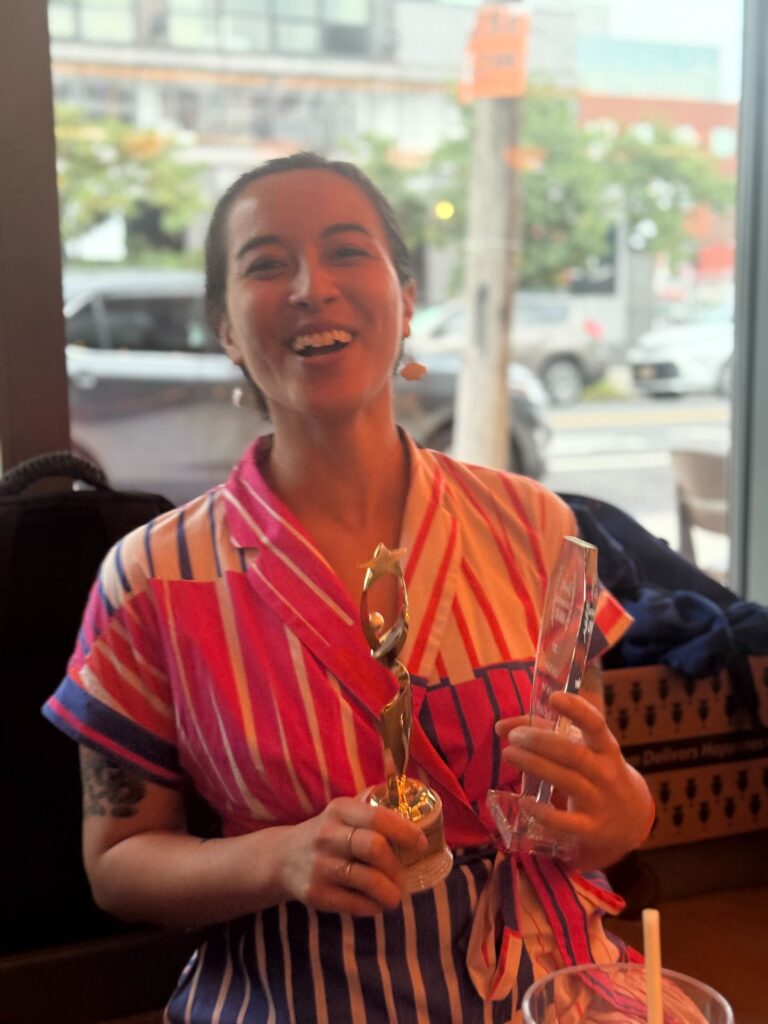 A woman holds two awards, one gold and the other clear. She is wearing a multi striped pink and white dress. 