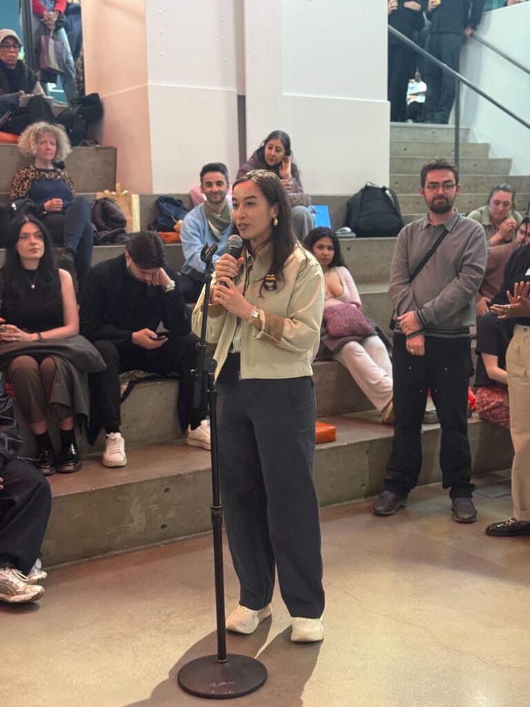 Woman speaks into a microphone at a public event, standing at the mic in front of a seated audience on steps.