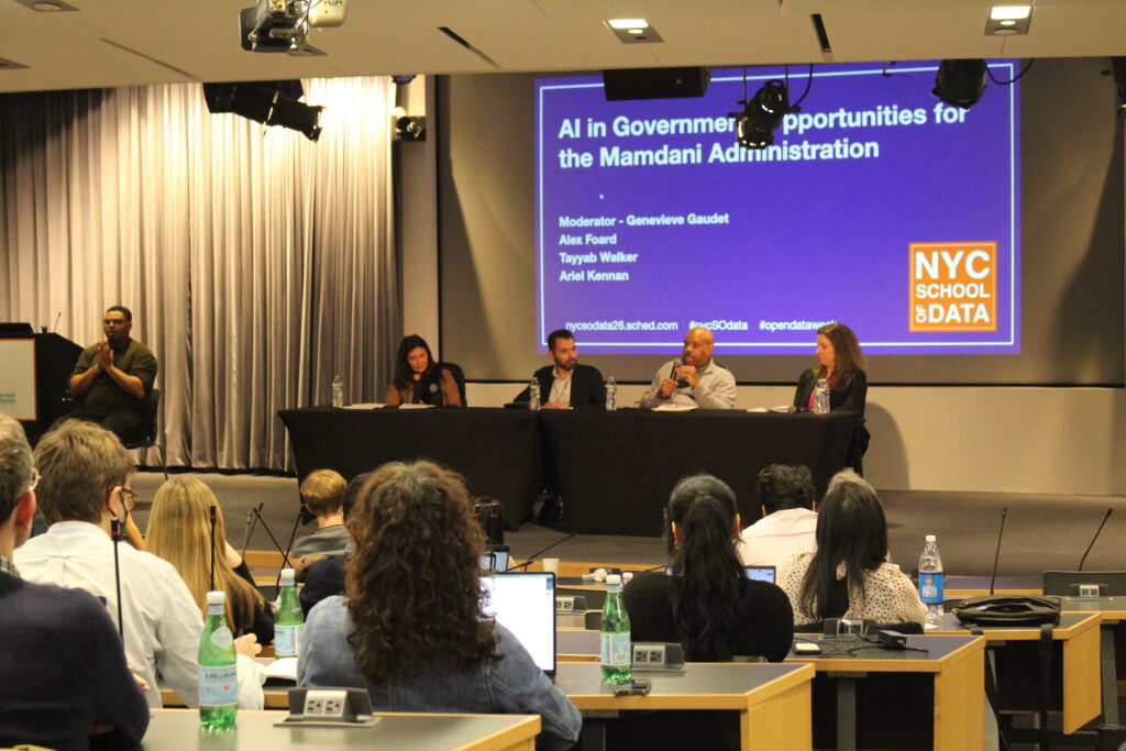 A wide-angle shot of a stage in a presentation hall. A panel of four diverse individuals is seated at a long, black-covered table. A projection screen behind them displays the title: "AI in Government: Opportunities for the Mamdani Administration," listing speaker names: Alex Foard, Ariel Kennan, Tayyab Walker, and Genevieve Gaudet