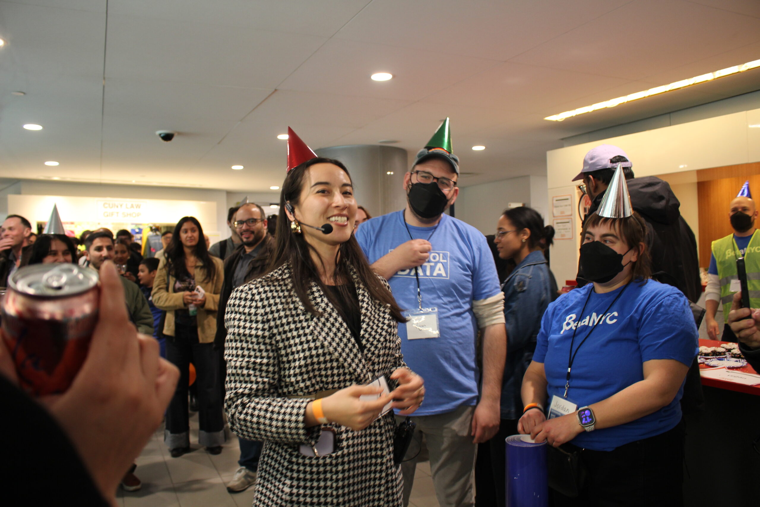 Jazzy Smith smiles in a plaid blazer and a red party hat, holding a microphone, while others around her wear different party hats.