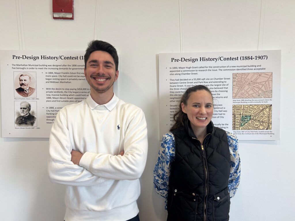 Two people stand next to each other. Behind them is a white wall and two images titled “Pre-Design History/Contest (1884-1907).