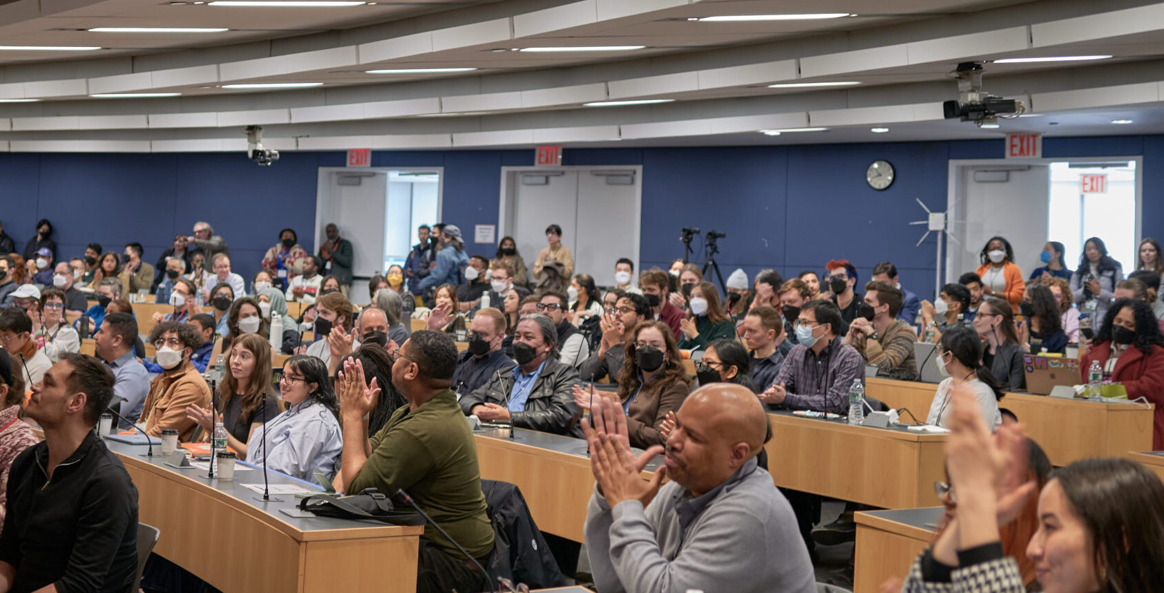 Large lecture hall filled with a diverse audience clapping during a conference.