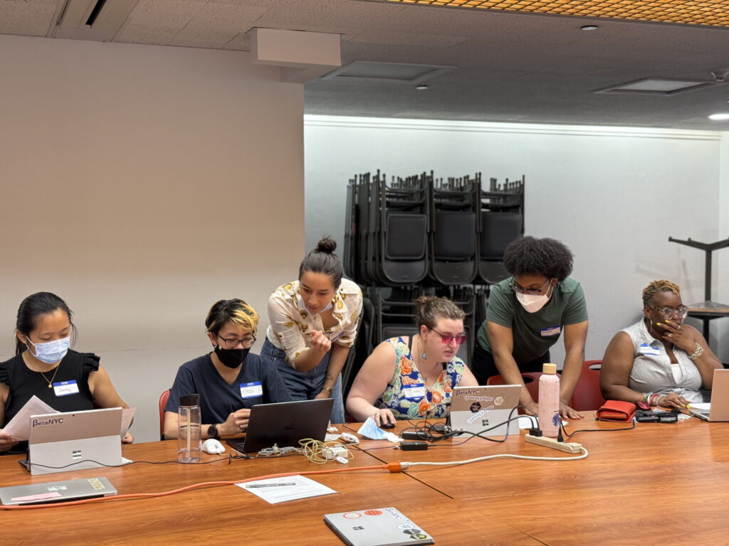Six people sit side by side at a long table covered with laptops, wires, and notebooks during a BetaNYC data entry workshop, while two facilitators stand behind them, leaning in to offer one-on-one help.