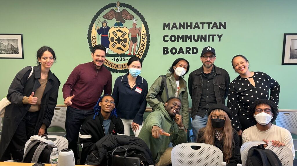 A group of participants pose inside a Manhattan Community Board meeting room, standing and sitting in front of a wall with the Manhattan seal and the words “MANHATTAN COMMUNITY BOARD,” some giving thumbs-up and several wearing masks.