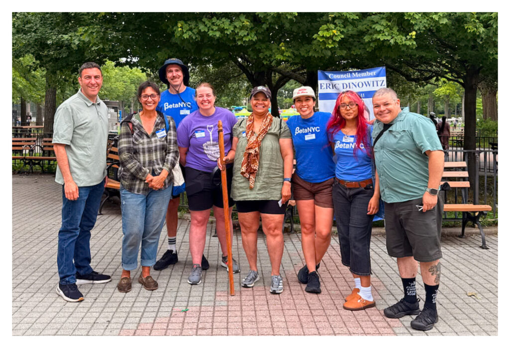 Eight people stand shoulder to shoulder in a paved park plaza with wooden benches and green trees behind them; several wear BetaNYC shirts and name tags, and a banner in the background reads “Council Member Eric Dinowitz.”