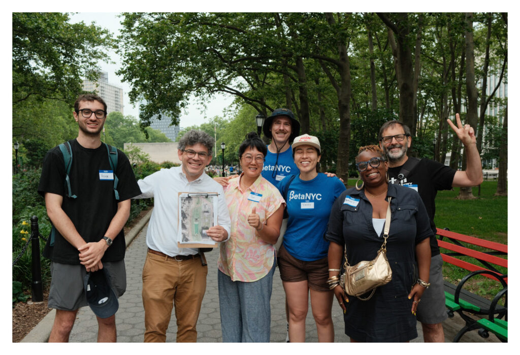 Seven people stand on a park path lined with trees and benches, smiling at the camera; one person holds a clipboard showing an aerial map, while two others wear blue BetaNYC shirts, and another flashes a peace sign.