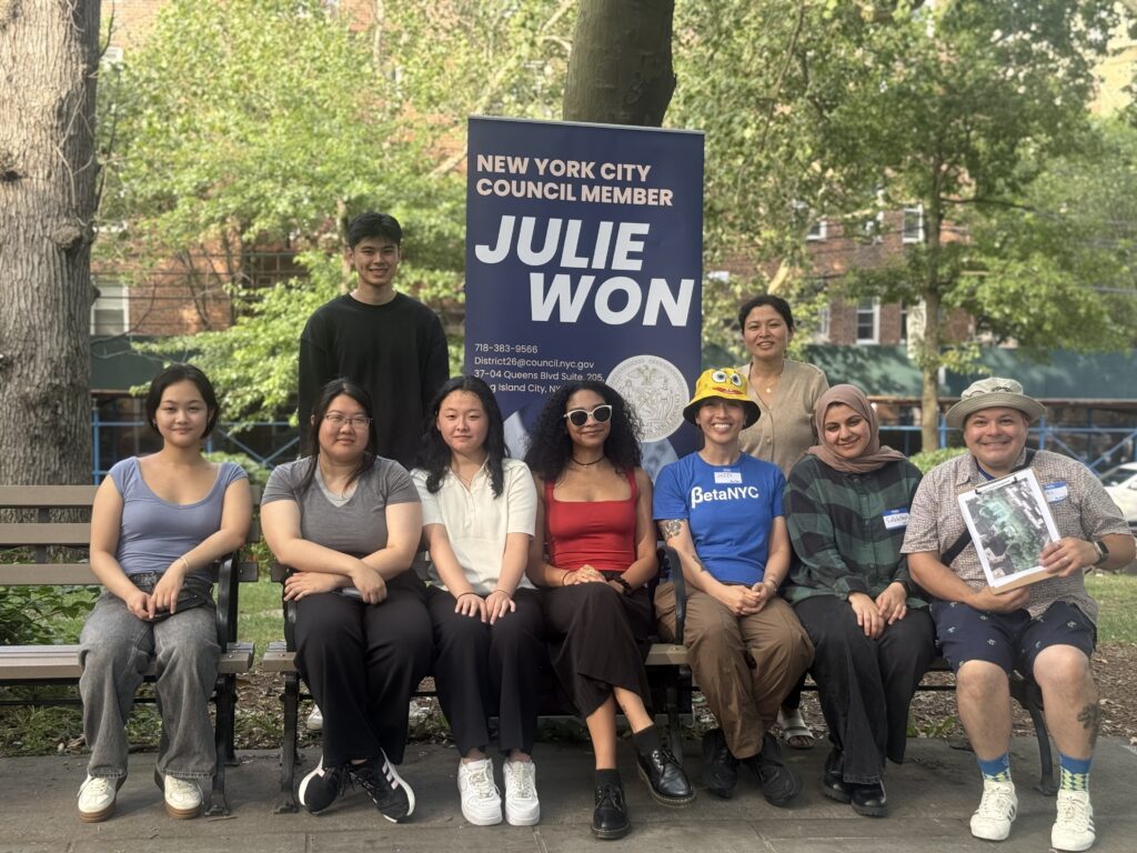 Nine people pose on and behind a park bench in front of a vertical banner that reads “New York City Council Member Julie Won,” with green trees behind them; several participants wear name tags, and one person in a BetaNYC shirt sits near the center.