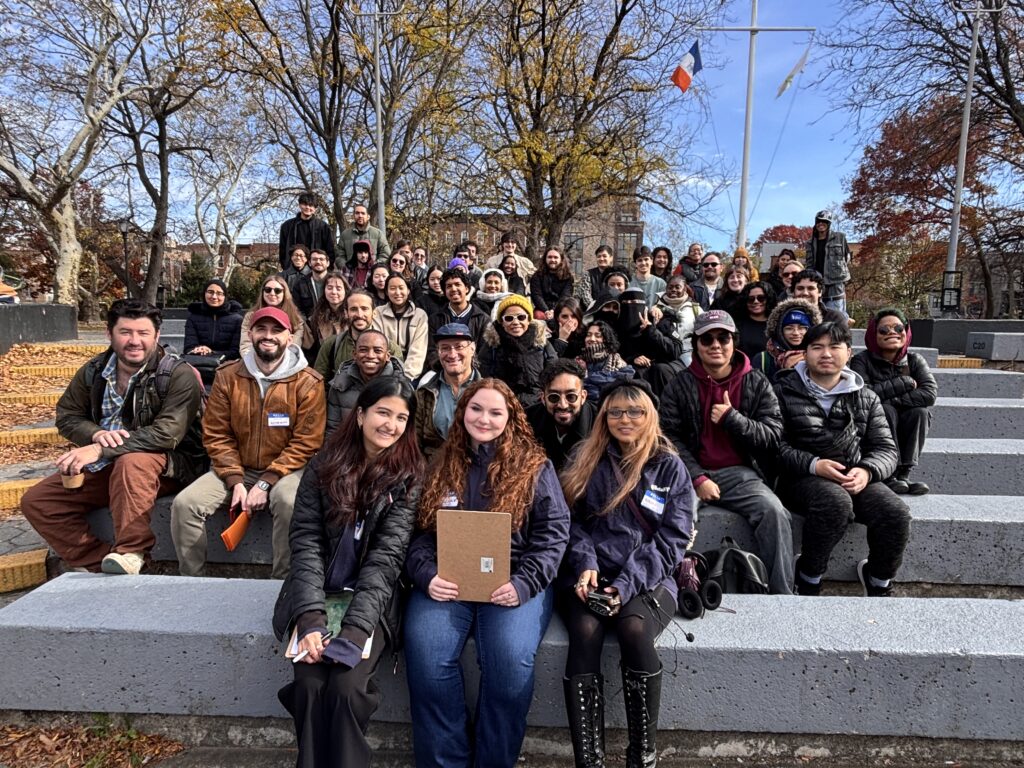 A large Mapping for Equity group sits closely together on outdoor concrete steps in a park on a crisp fall day, bundled in jackets and hats and smiling at the camera, with bare trees and a flagpole in the background.