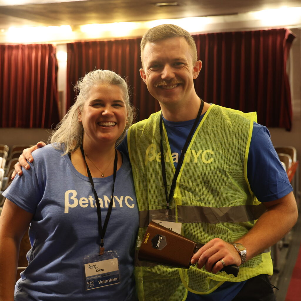 Two volunteers in BetaNYC shirts, one in a safety vest, smile in the auditorium aisle.