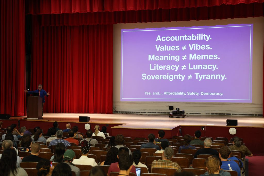 Noel Hidalgo speaks from a podium in the Hunter College auditorium beside a slide reading "Values ≠ Vibes. Meaning ≠ Memes. Literacy ≠ Lunacy. Sovereignty ≠ Tyranny."