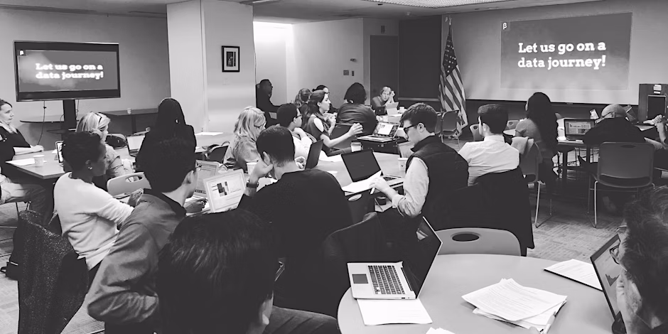 A group of people sit at various tables with laptops, surrounded by monitors.