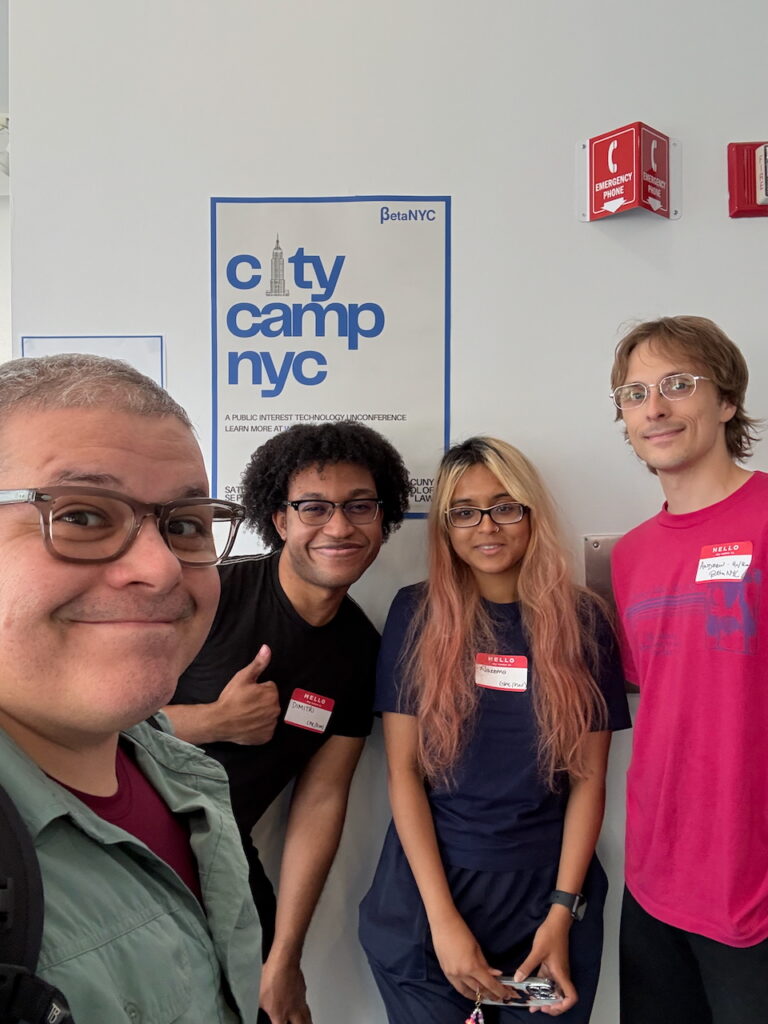BetaNYC staff stand together in a hallway in front of a CityCamp NYC poster, smiling at the camera with conference name tags on their shirts.