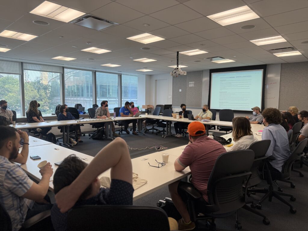 Participants sit around a large rectangular table in a bright conference room, with two facilitators at the front leading a CityCamp NYC discussion and a shared document projected onto a screen.