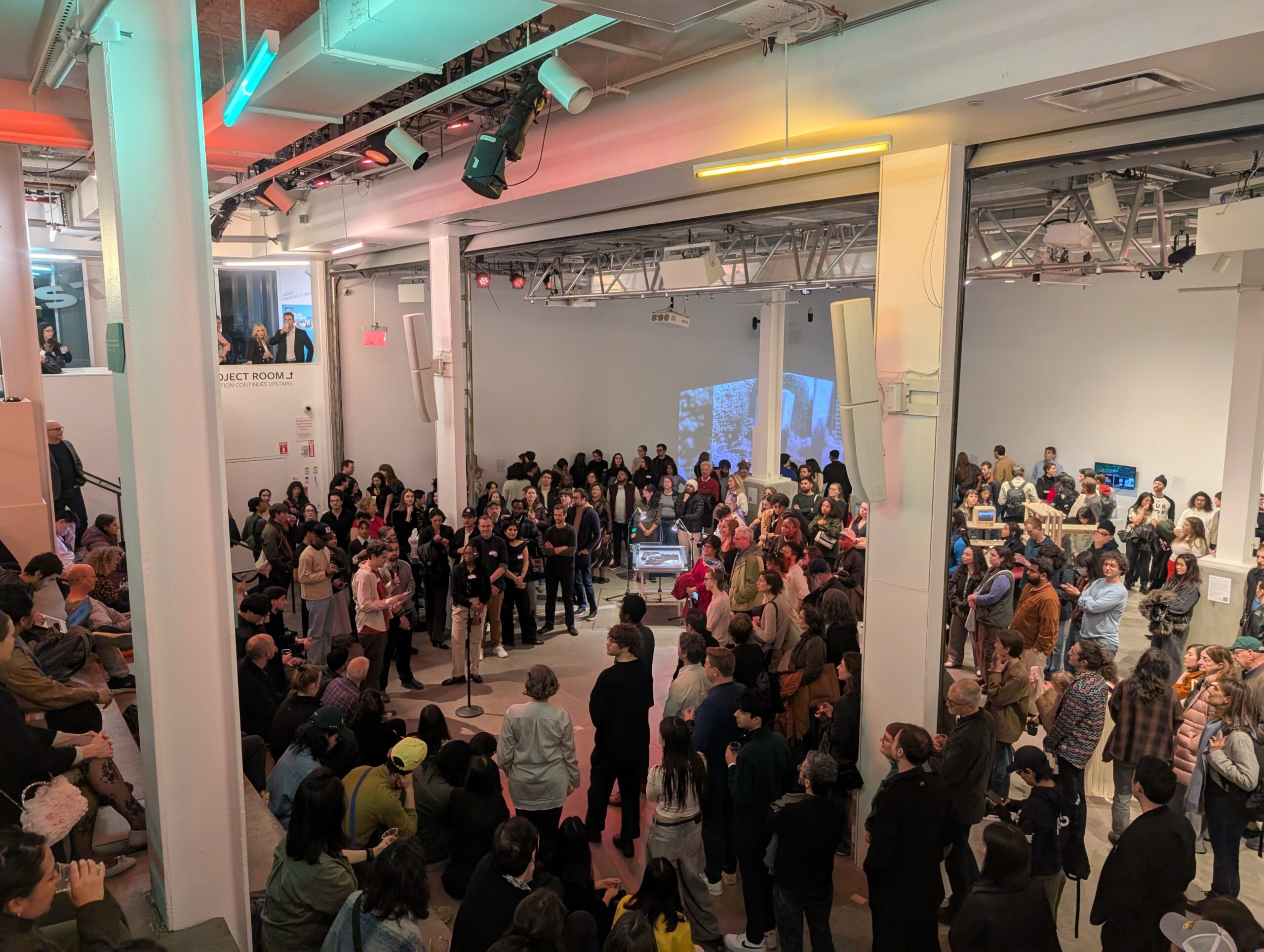 A large crowd of people gathers in a spacious room with a space for opening remarks at the Data Through Design exhibition opening event at BRIC
