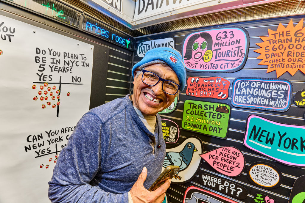 Smiling man with glasses and a blue beanie posing in front of a wall of colorful NYC-themed signs and graphics, holding a phone.
