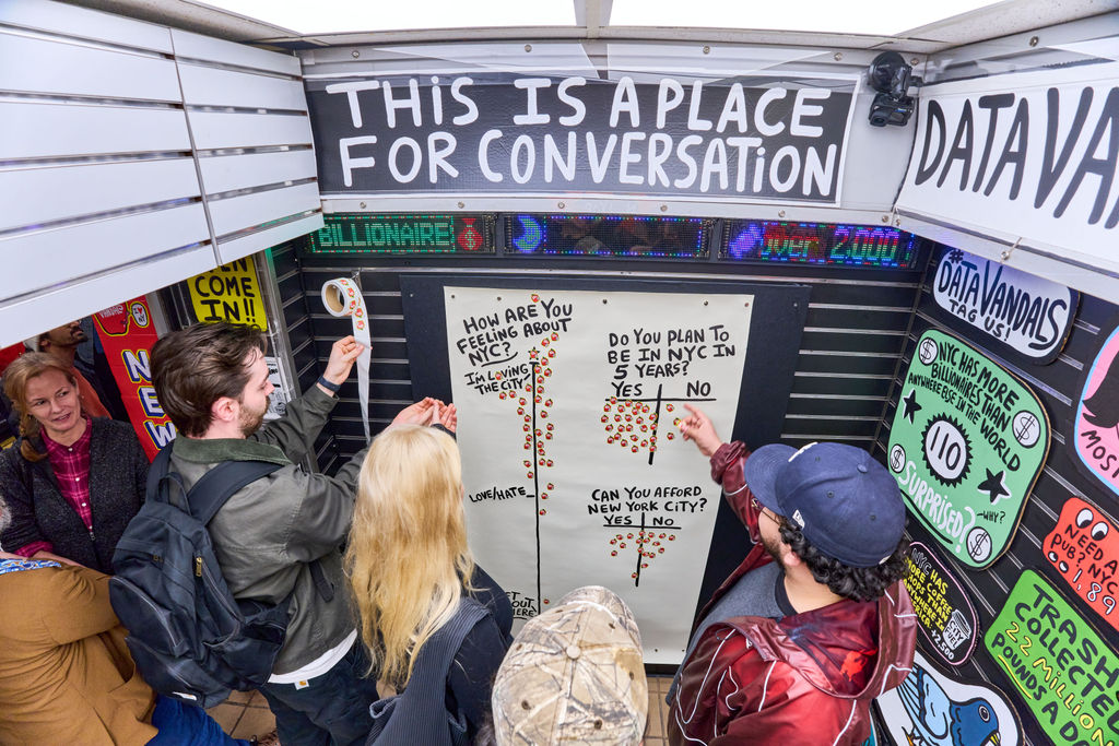 People in a small booth pin sticky notes to a large whiteboard while others watch, under a banner that reads 'This is a place for conversation.'