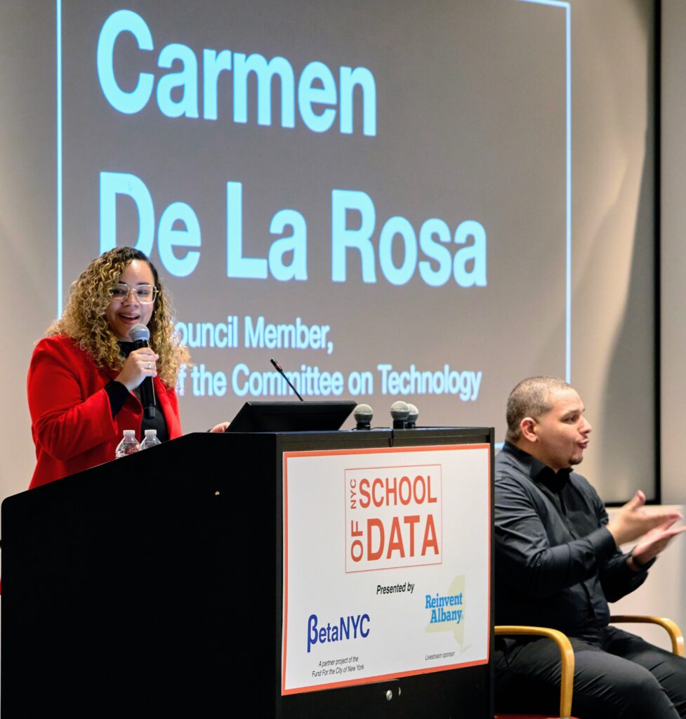 NYC Council Member Carmen De La Rosa smiling and speaking into a microphone while standing in front of a NYC School of Data lectern