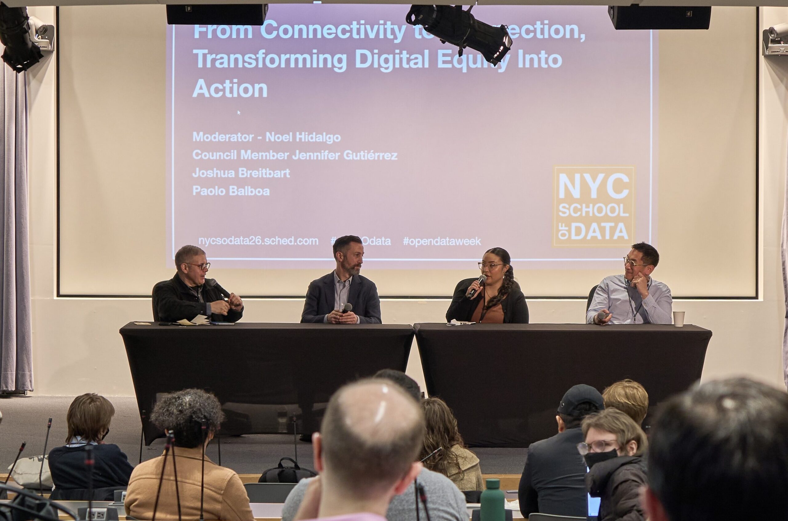 A photograph of a four-person panel seated behind black table coverings on a stage. The panel members are seated from left to right: Noel Hidalgo, Joshua Breitbart, Council Member Jennifer Gutiérrez, and Paolo Balboa. Behind them is a large projection screen displaying the text "From Connectivity to Connection, Transforming Digital Equity Into Action"