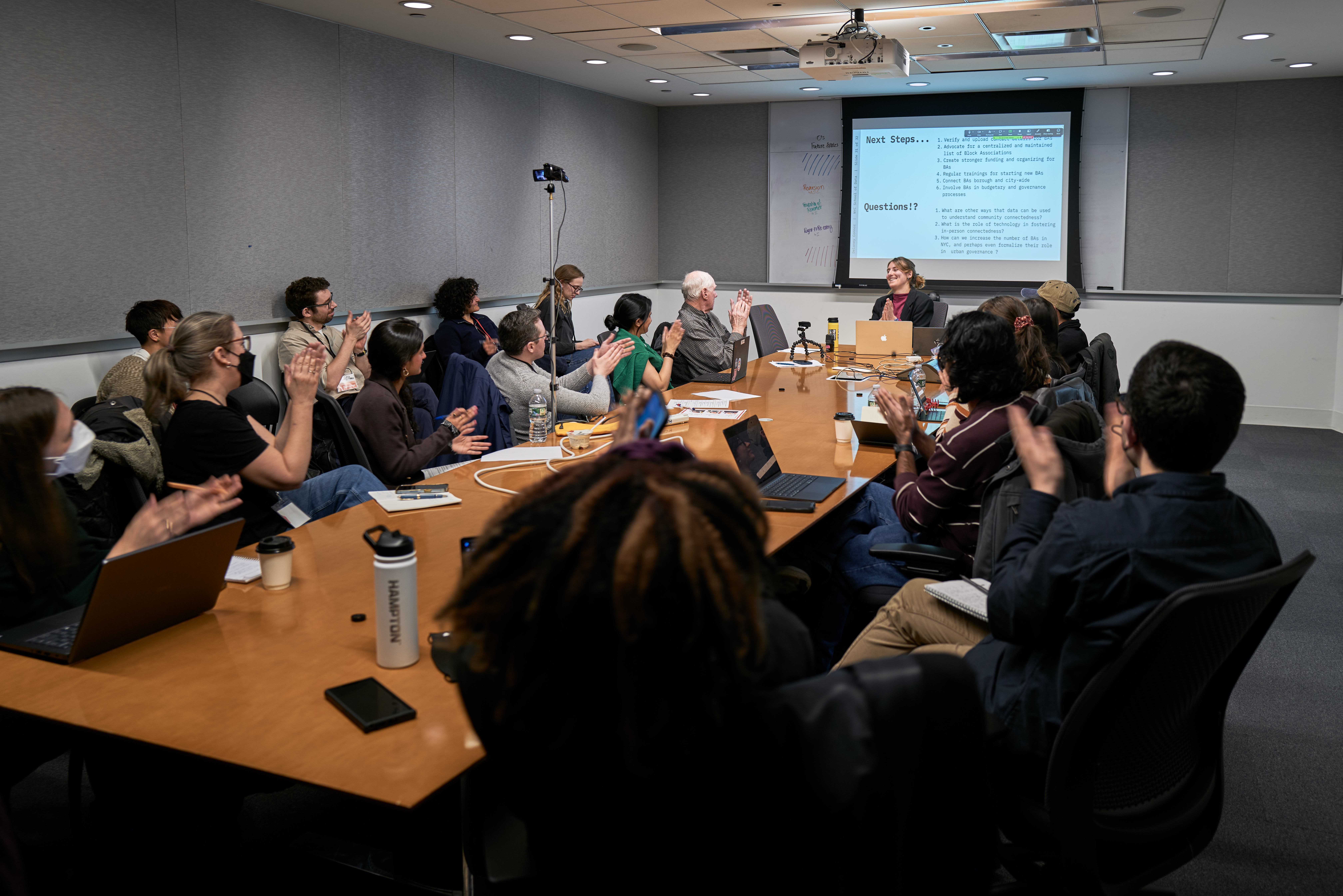 A diverse group of about 15 people are seated around a long wooden conference table. Most are looking forward toward a presenter and clapping their hands. The room is a conference space with a projector screen at the far end showing a "Next Steps..." checklist.