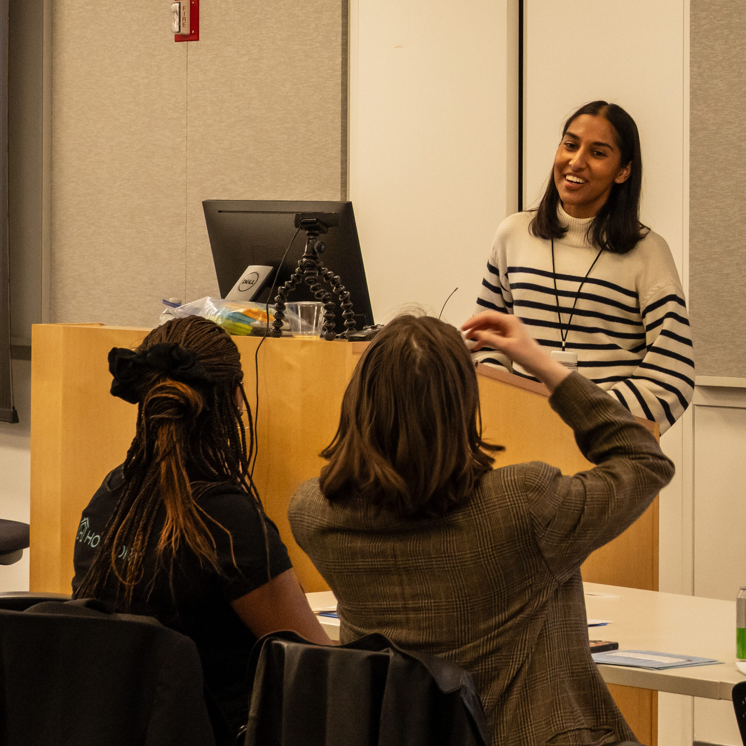 A young woman in a striped sweater smiles as she leads a presentation from a lectern. Two people in the foreground listen from their seats.