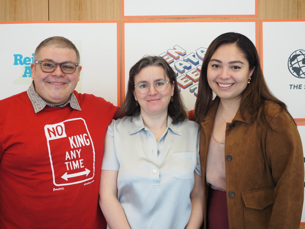 Three people stand close together and smile at the camera in front of a colorful sponsor backdrop; one wears a red “No King Any Time” T-shirt, and the others wear business-casual tops and jackets.