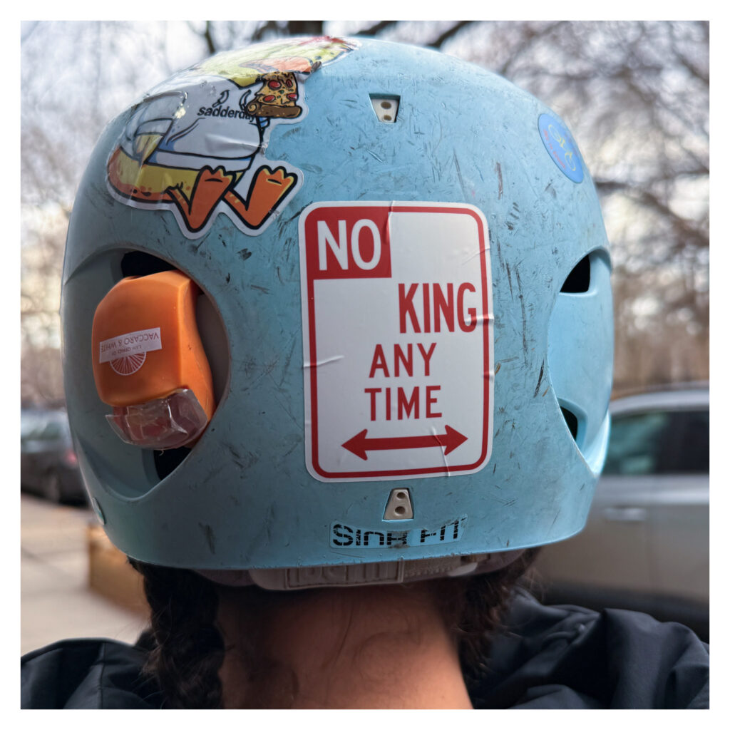 A person wears a light blue helmet with various stickers, including a red and white sticker that says "NO KING ANY TIME."