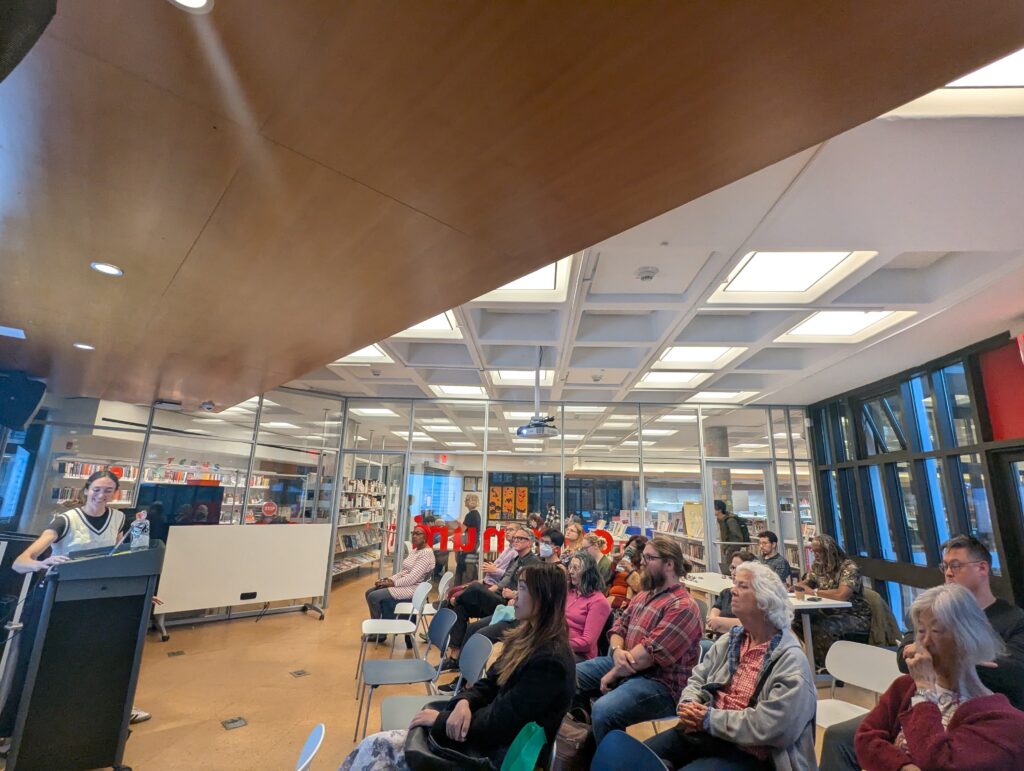 Wide view of a glass-walled library room during a talk: a speaker stands at a lectern on the left as an audience of adults listens, with bookshelves, ceiling lights, and large windows in the background.