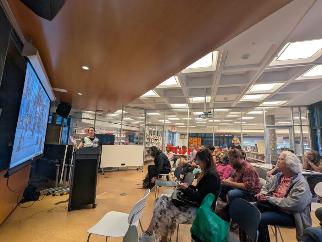 A presenter at a lectern speaks beside a projected photo of a busy Union Square street scene; participants sit in rows, some taking notes or checking phones, in a bright library space.