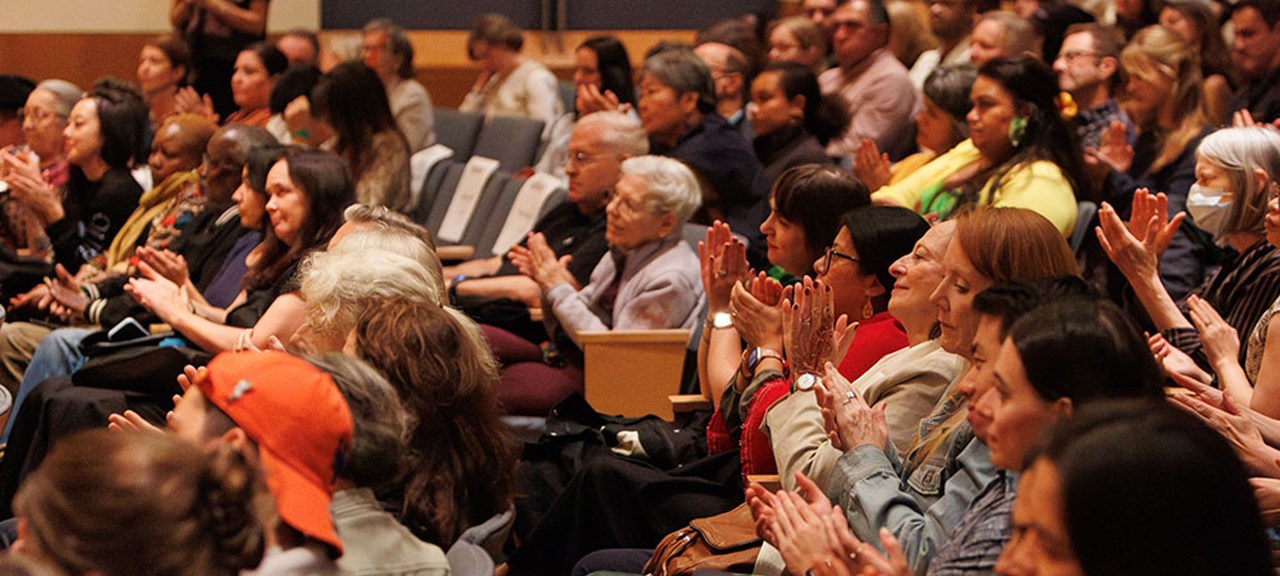 Audience seated in an auditorium applauding; diverse group of men and women of various ages watching a performance.
