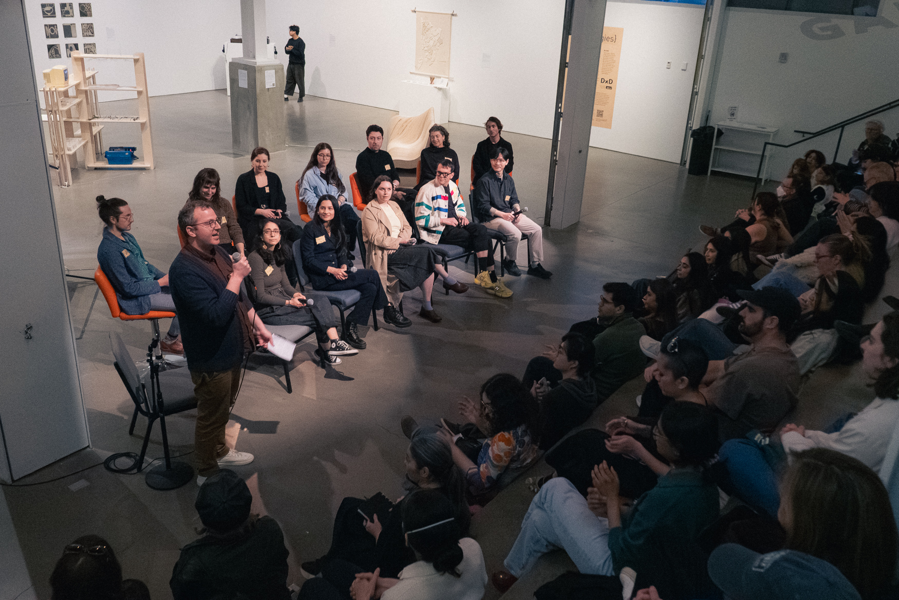 A wide shot of a panel discussion taking place in a large, modern industrial space at BRIC. On the left, about a dozen Data Through Design Artists sit in a semi-circle on orange and blue chairs. A man in a dark cardigan and tan pants stands in front of them, speaking into a microphone.
On the right, a large audience is seated on tiered bleacher-style seating, listening attentively.