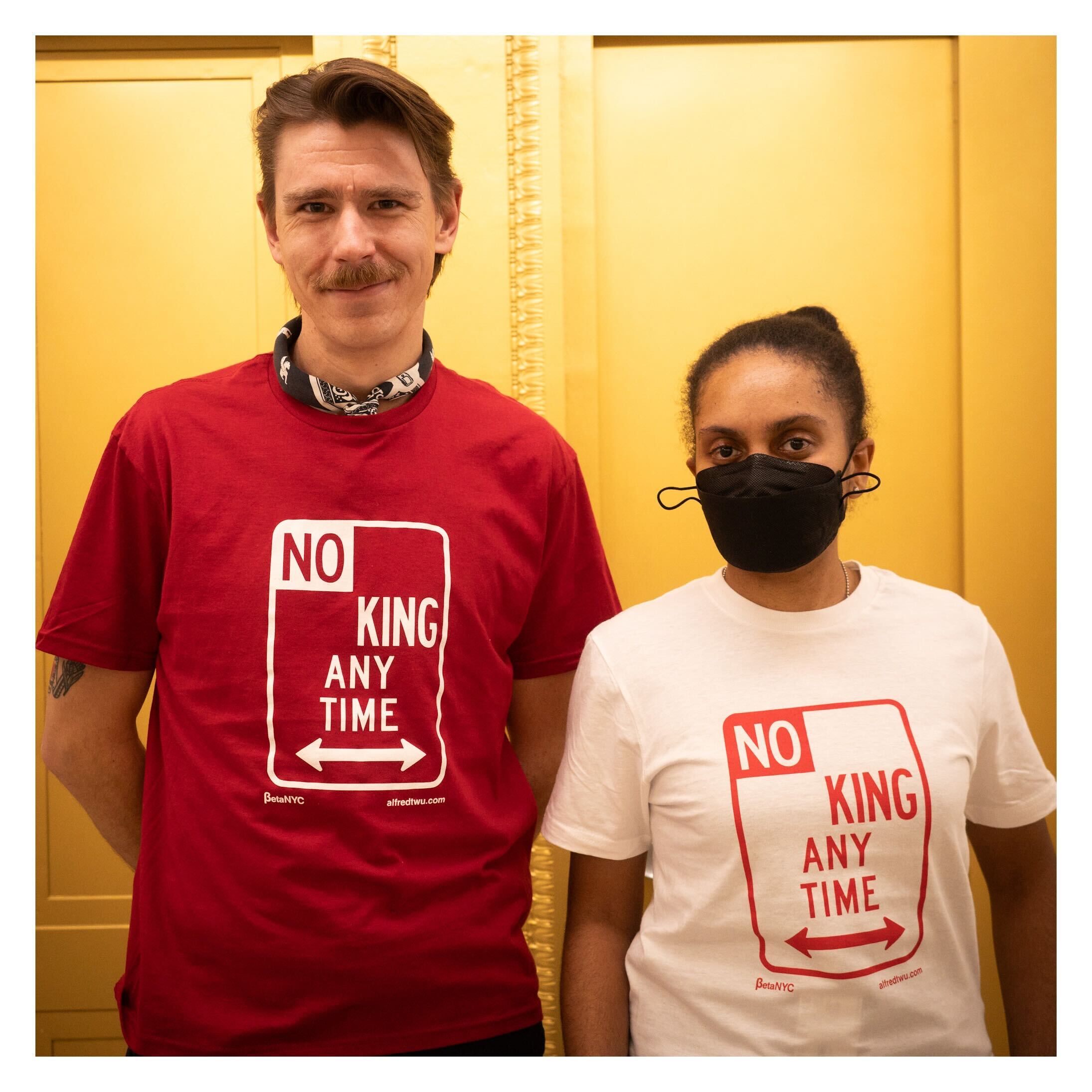 Two people stand side by side in front of a gold wall. The person on the left is wearing a red T-shirt with white text that reads “NO KING ANY TIME,” designed in the style of a New York City parking sign. The person on the right is wearing the same shirt design in white with red text and is also wearing a black face mask. Both shirts feature logos for BetaNYC and alfredtwu.com at the bottom.