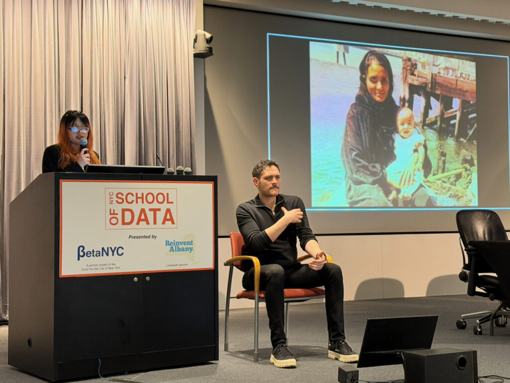 A woman speaks at a podium containing a sign with the logos for School of Data, BetaNYC, and Reinvent Albany. Next to her on stage is an ASL interpreter and a large image of a woman holding a child.