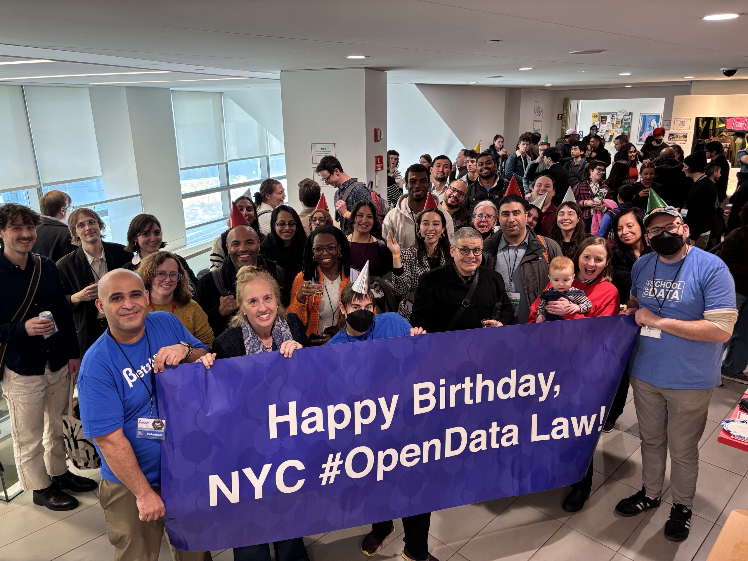A large group of people stand in a cafe/eating space. The row of people in the front are carrying a dark blue banner that reads: "Happy Birthday, NYC #OpenData Law!"