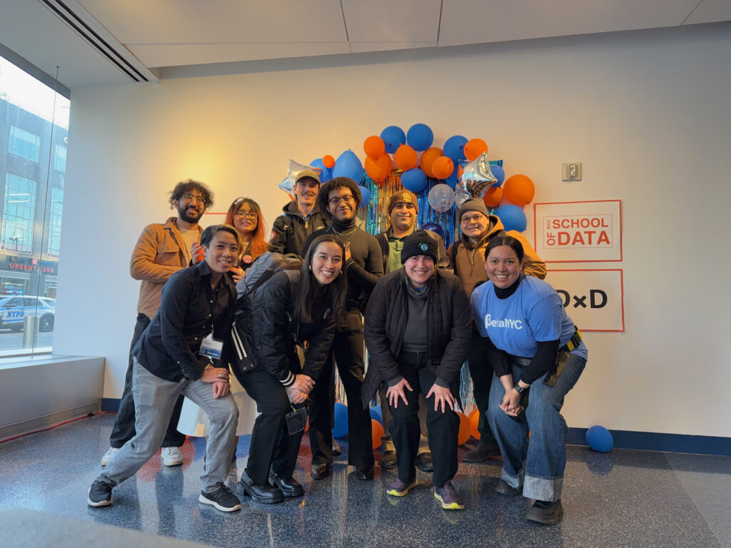 BetaNYC staff and volunteers pose in front of a white wall with blue and orange balloons and various signage.