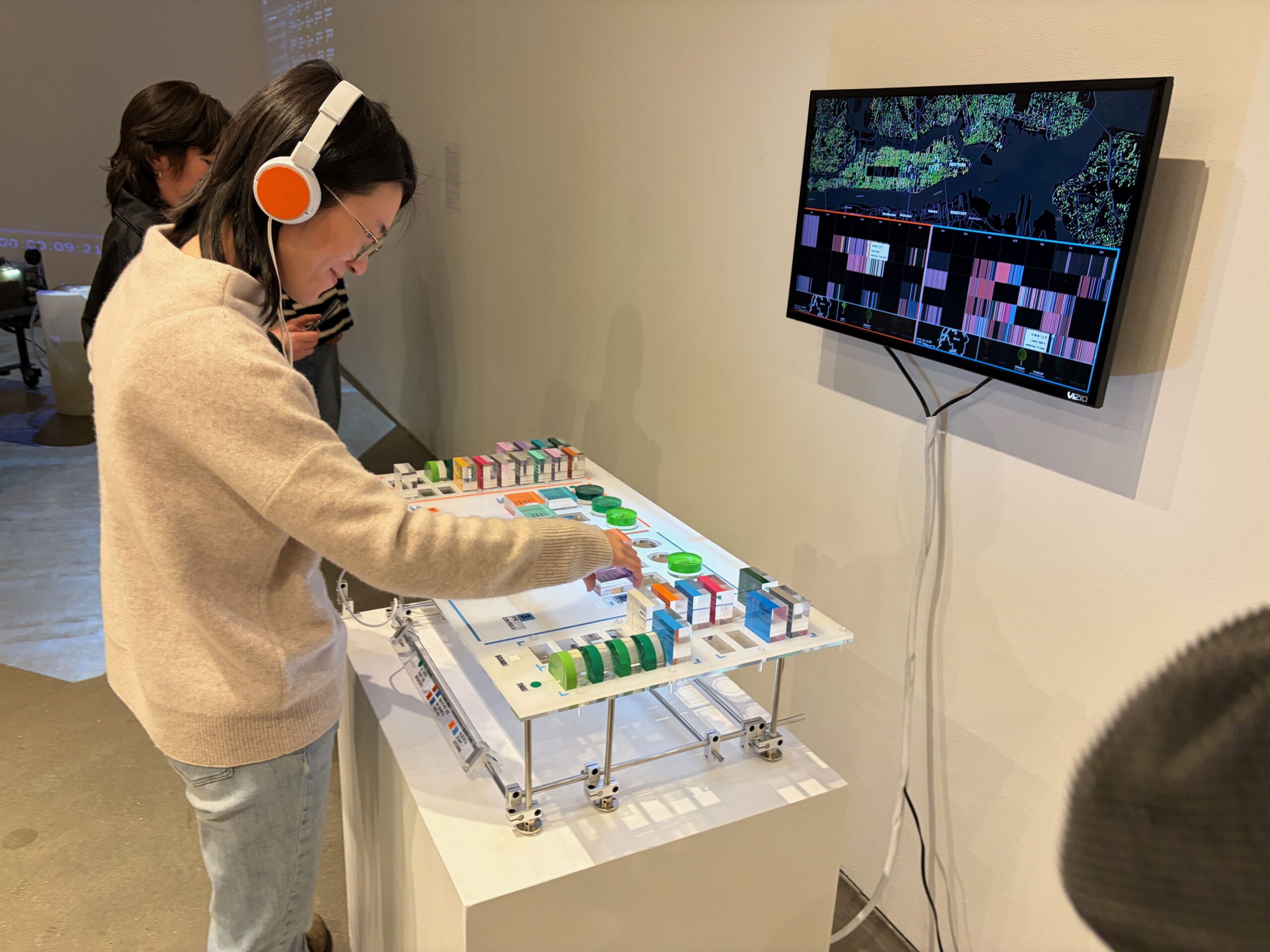 An individual with a smile is fully engaged in a installation game featuring green and red blocks. They wear headphones while the screen in the background displays data relevant to the gameplay.